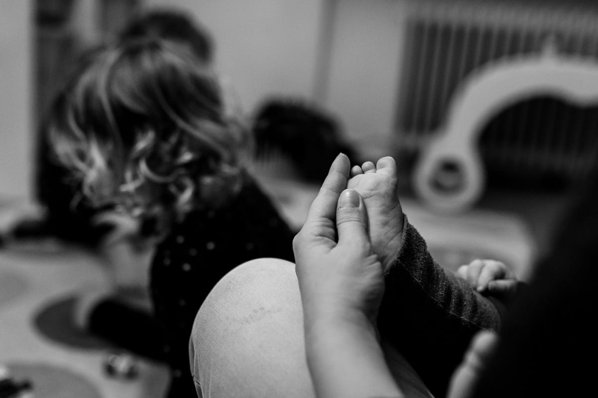 Hand einer Mutter fasst zärtlich ein Babyfuß an, im Hintergrund spielt ein kleines Mädchen.