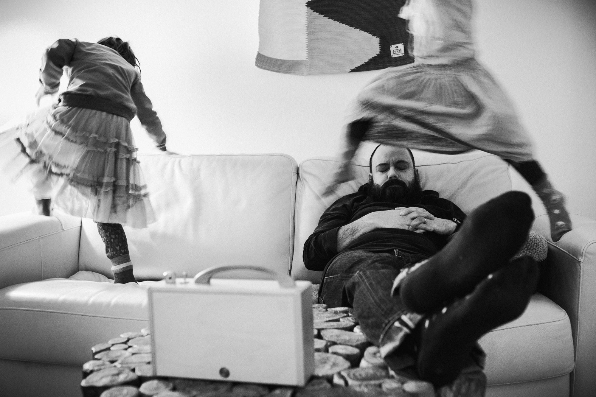 Ein Vater schläft auf der Couch. Kinder springen schnell um ihn herum.