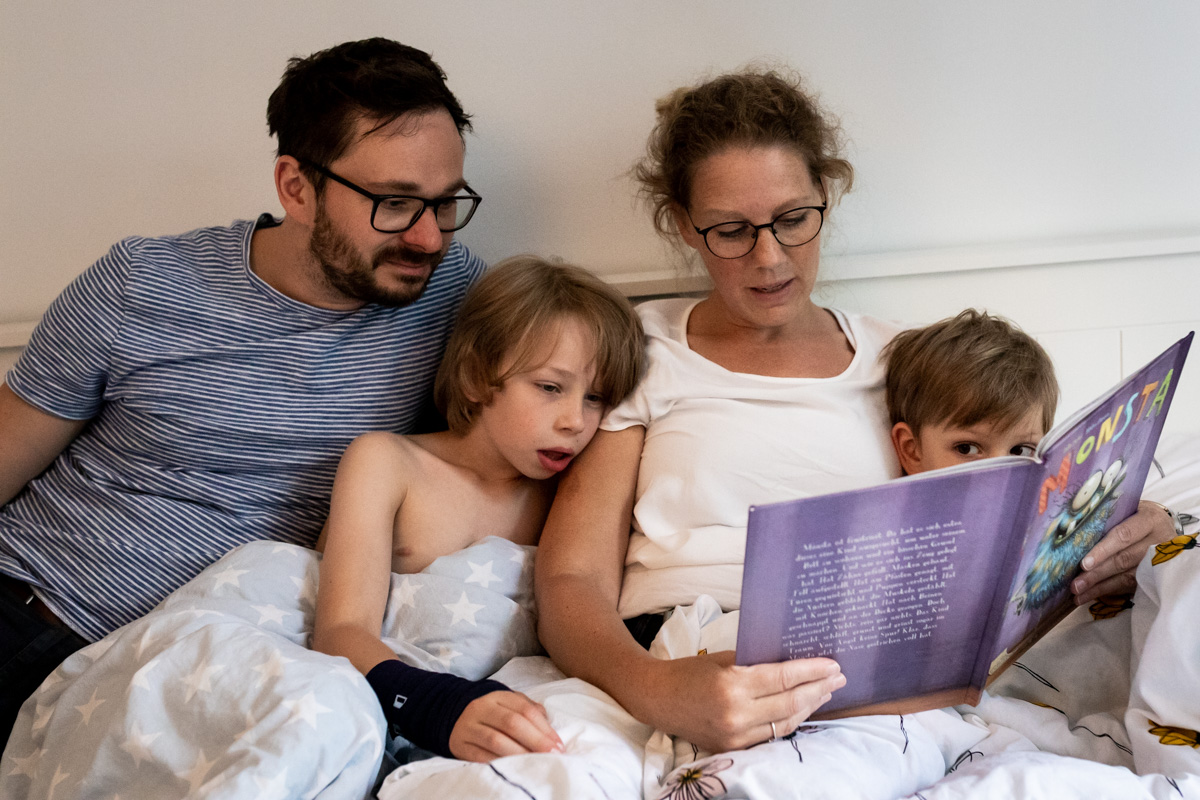 Eltern mit zwei Kindern im Bett. Es wird ein Buch vorgelesen.