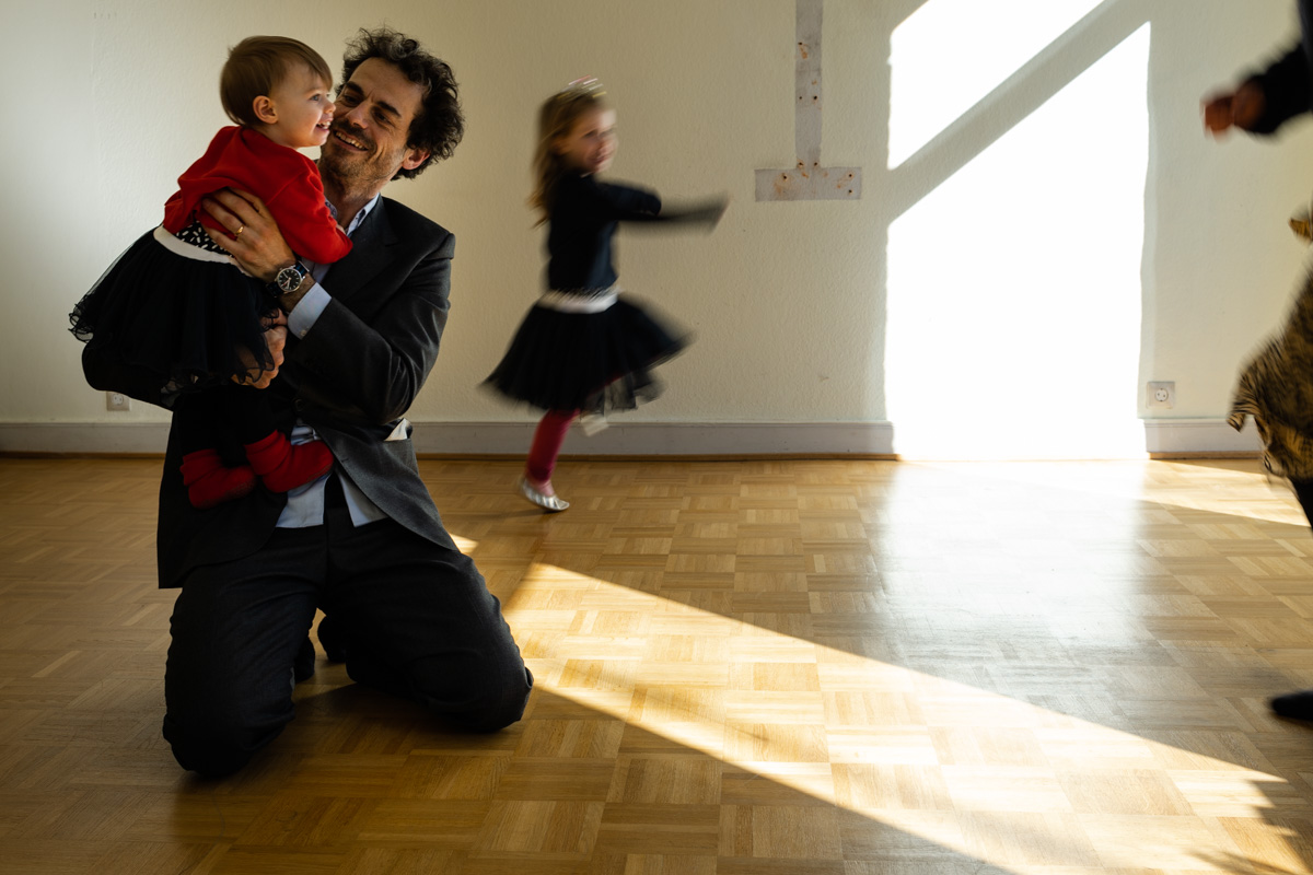 Ein Vater kniet und hält seine Tochter auf dem Arm. Drumherum tanzt seine größere Tochter. Man sieht sie in Bewegung.